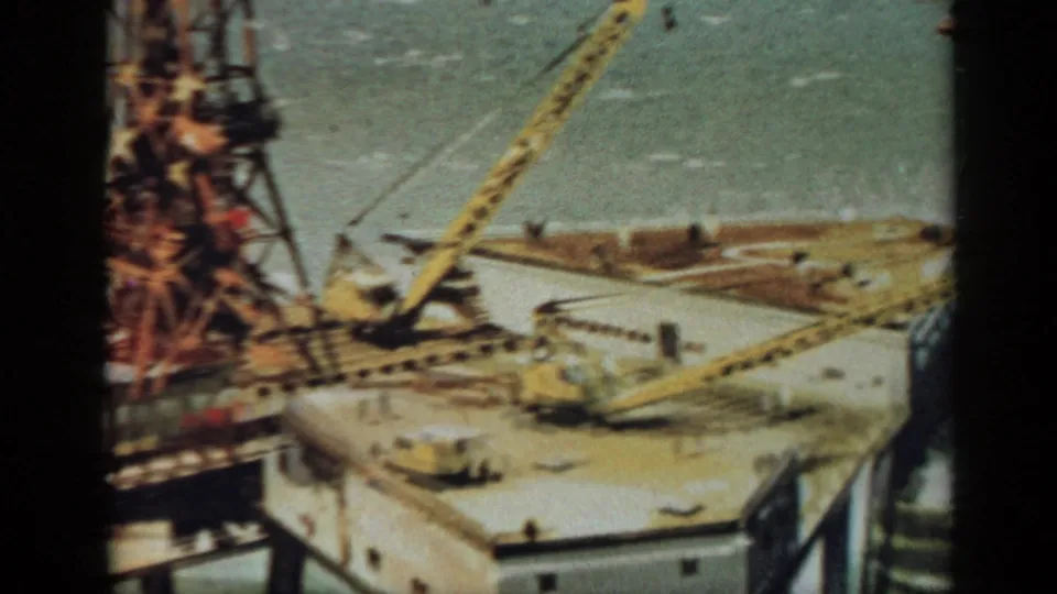 1970 United Kingdom 8mm frame with a closer view across an offshore platform, showing crane booms, a central tower, deck structures, and water.
