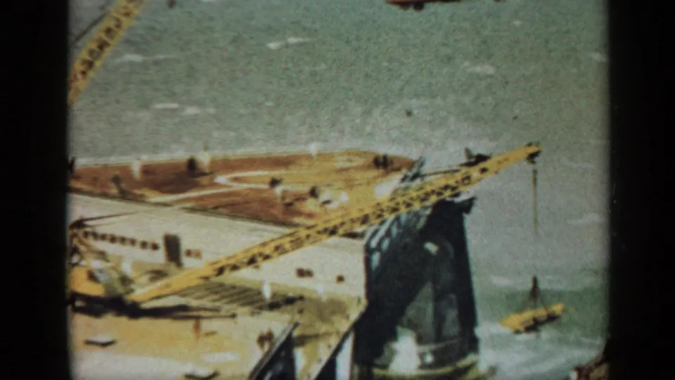 1970 United Kingdom 8mm frame showing a long yellow crane over an offshore platform corner, with deck structures, distant workers, and surrounding water.