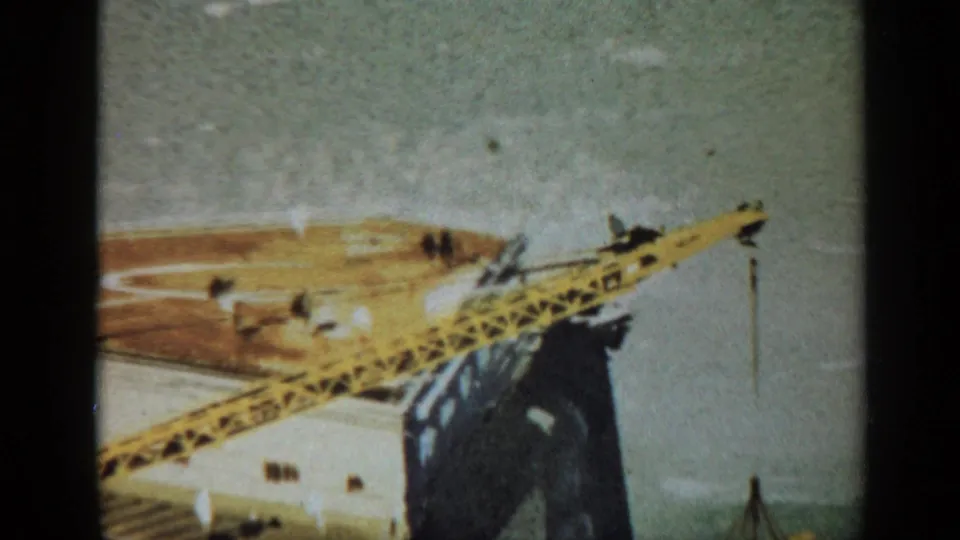 1970 United Kingdom 8mm frame showing the edge of an offshore platform with a yellow crane, deck activity, small workers, and open water behind.