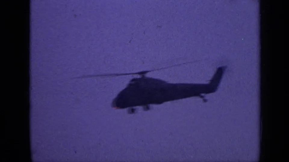 1970 United Kingdom 8mm frame of a helicopter in silhouette, with rotor blur against a purple-blue sky.