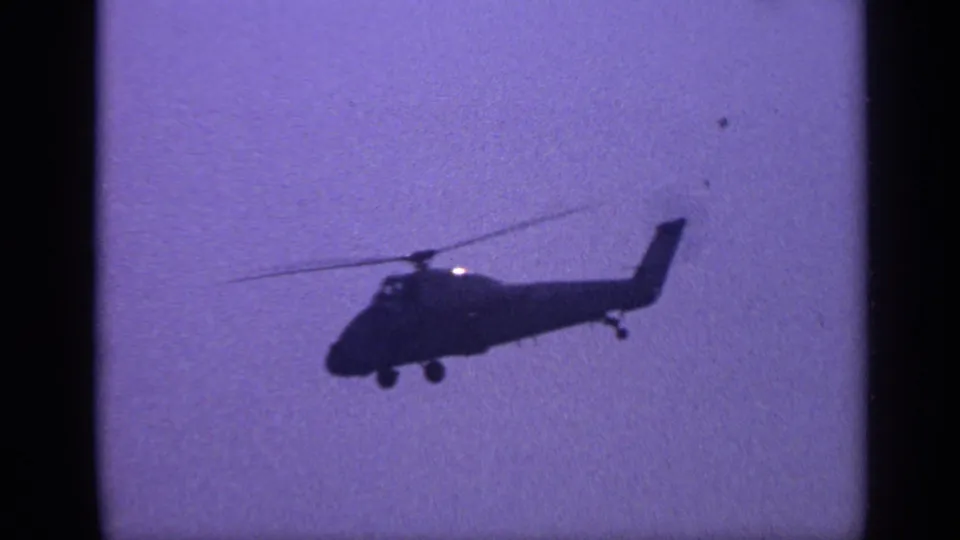 1970 United Kingdom 8mm frame showing a silhouetted helicopter in profile against a hazy, purple-tinged sky.