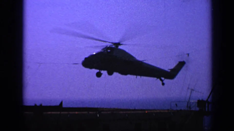 1970 United Kingdom 8mm frame of a military helicopter in profile, rotor blurred, descending near a ship deck at twilight.