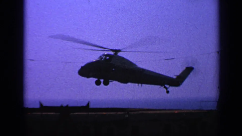 1970 United Kingdom 8mm frame showing a helicopter passing low over the ship area, with rotor blur and landing gear visible.