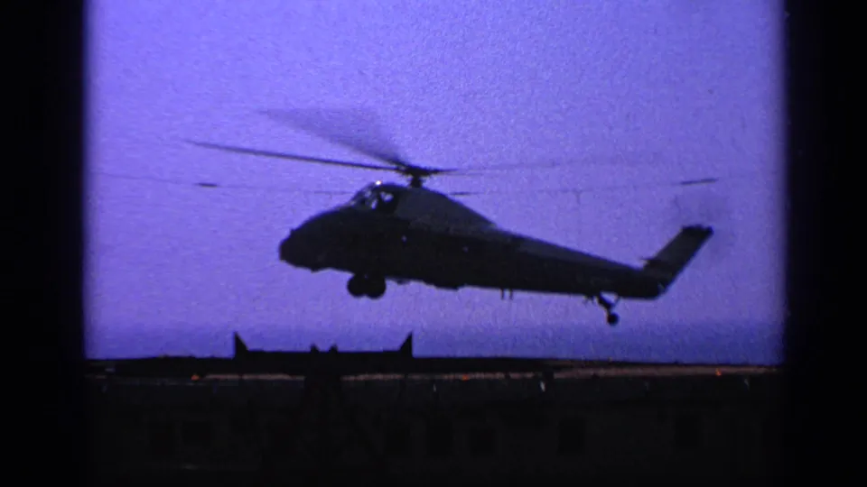 1970 United Kingdom 8mm frame of a helicopter nearing the ship deck, seen in silhouette with landing gear down and rotor motion blur.