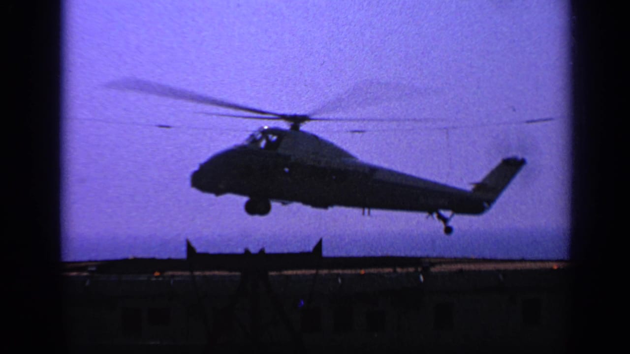 Silhouetted helicopter descending above a dark deck against a purple sky.