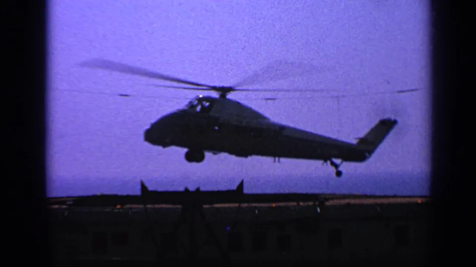 1970 United Kingdom 8mm frame showing a silhouetted military helicopter approaching a deck with landing gear extended.