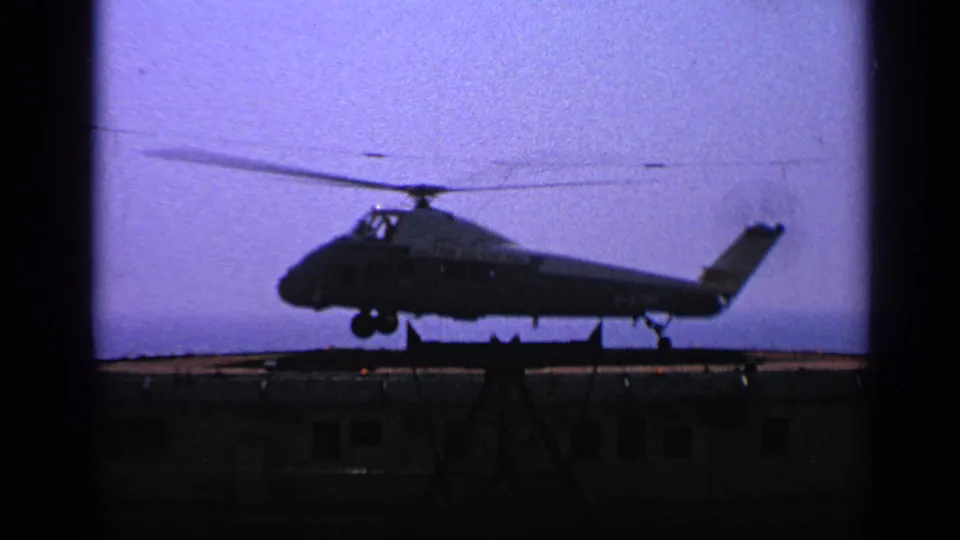 1970 United Kingdom 8mm frame of a silhouetted military helicopter completing a soft landing on a dark deck.