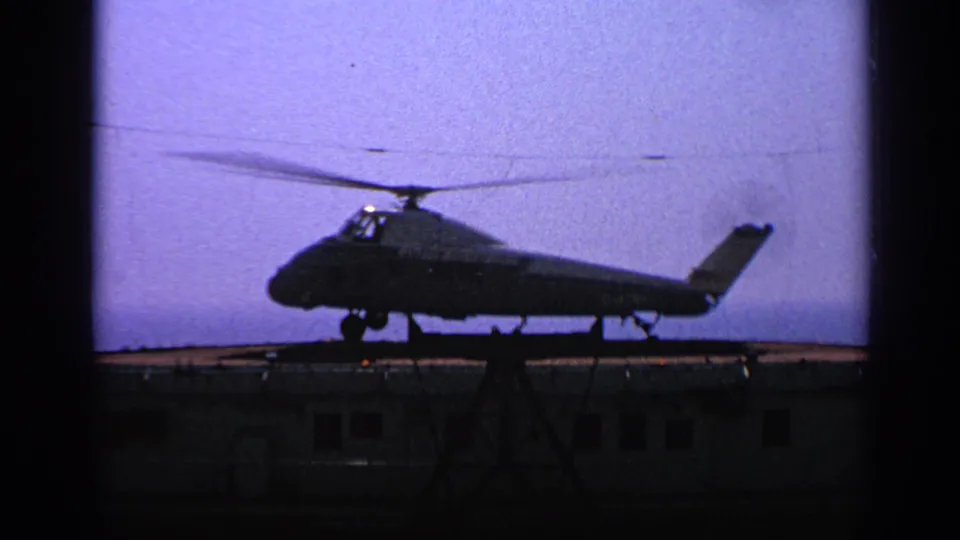 1970 United Kingdom 8mm frame showing a dark military helicopter silhouette on or near a small landing platform at twilight.