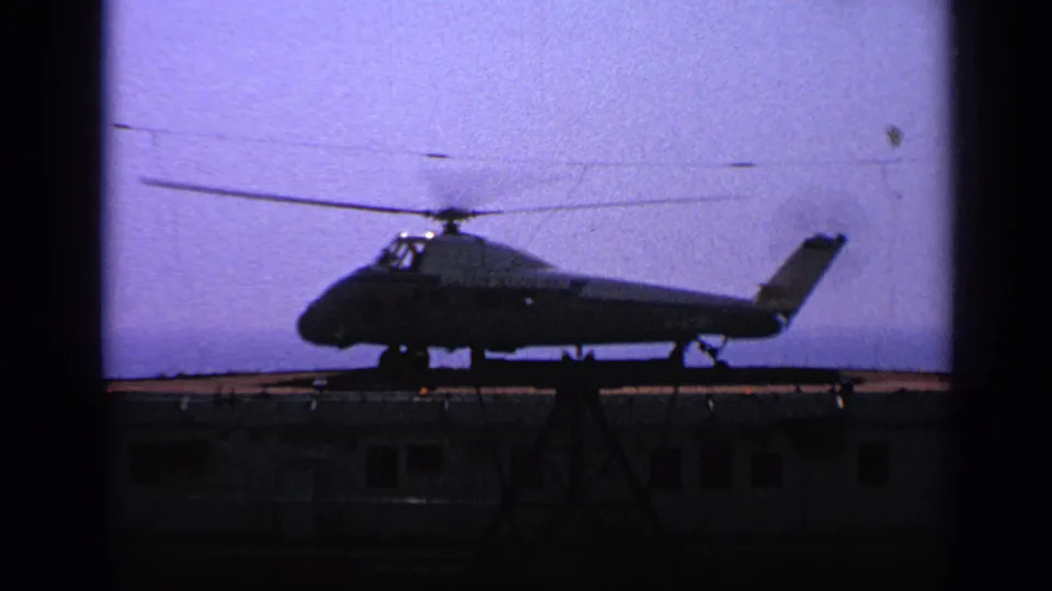 A dark helicopter profile appears on a small air pad in 1970 United Kingdom 8mm footage, with rotor blades spanning the sky.