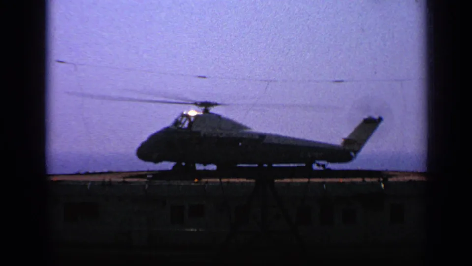 Final 1970 United Kingdom 8mm frame of the helicopter in silhouette on the small landing platform, with dark structures below.