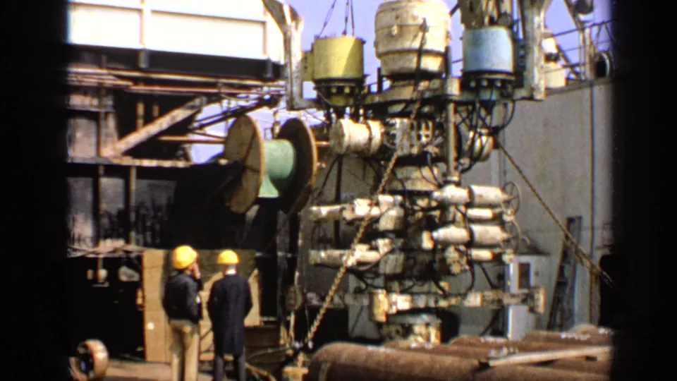 1970 United Kingdom 8mm frame with hard-hatted workers beside dense drilling machinery, pipes and rig structures on deck.
