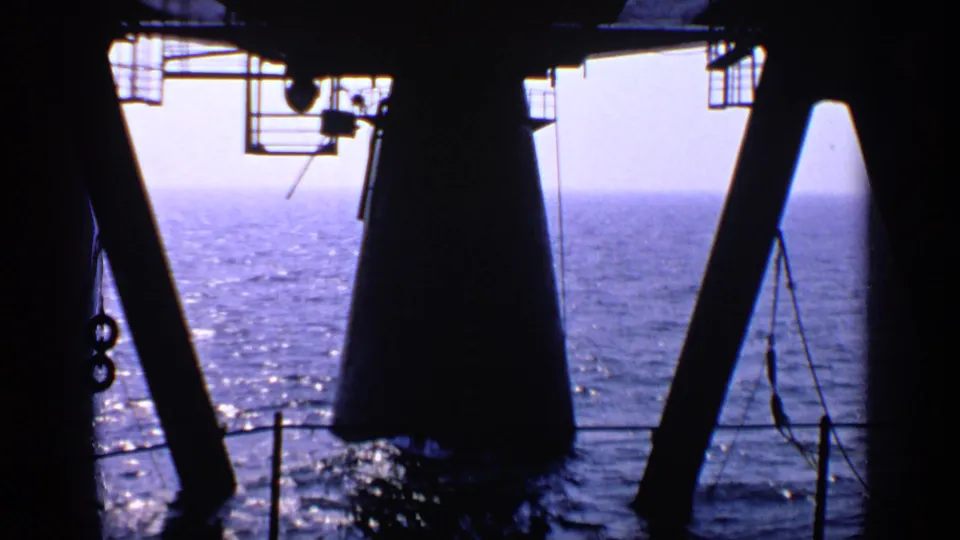View from an offshore rig structure toward open water, with silhouetted beams, railing, and equipment overhead.