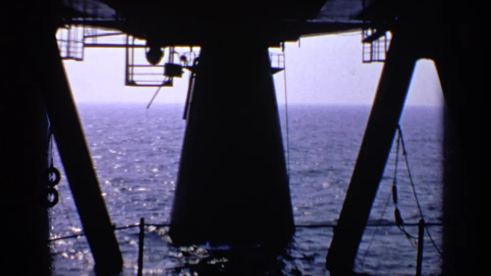 Offshore platform perspective looking past dark rig supports and railings across the ocean under a pale sky.