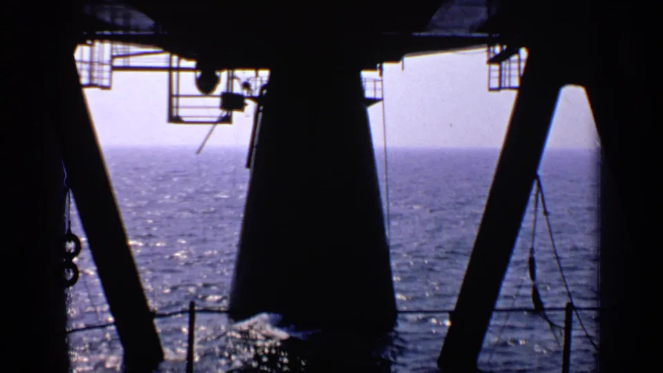 Silhouetted offshore rig structure with beams, railing, and suspended lines looking outward over the water.