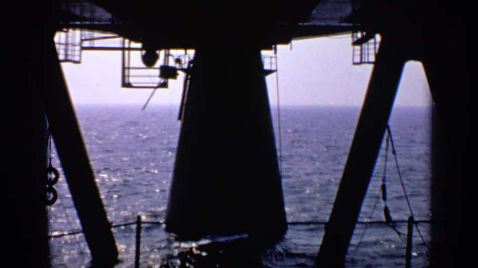 1970 United Kingdom 8mm frame showing dark ship or platform structures around sunlit open water and a hazy horizon.
