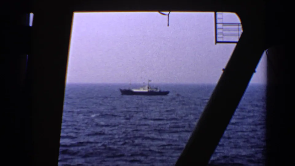 1970 United Kingdom 8mm frame showing a distant vessel on open water, viewed through the dark interior framing of a ship window.