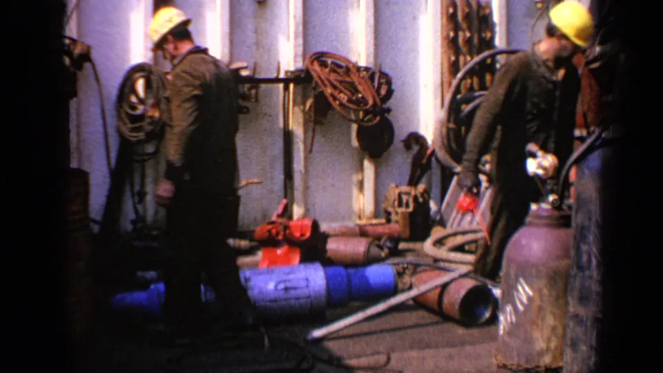 Two hard-hatted workers on an industrial deck handle equipment among hoses, pipes, cylinders, and fittings in 1970 United Kingdom footage.