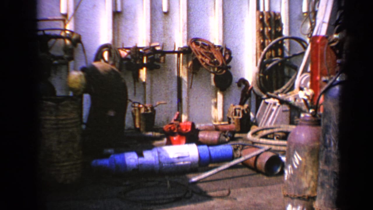 Worker bends near hoses and machinery inside a pipe-filled industrial area.