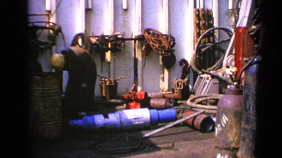A hard-hatted worker appears to handle equipment beside hoses, pipes, and valves in a 1970 United Kingdom industrial setting.