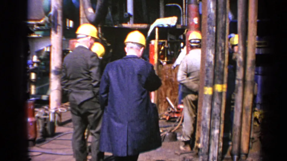 Several men in yellow hard hats and dark coats stand together amid machinery and vertical pipes at a 1970 United Kingdom industrial site.
