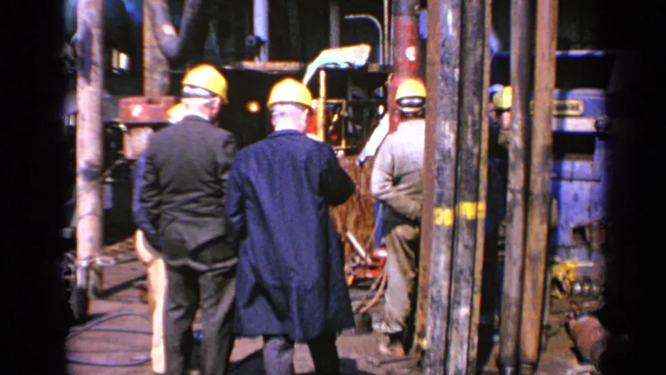 Men in yellow hard hats stand with their backs partly turned beside pipework and machinery in 1970 United Kingdom worksite footage.
