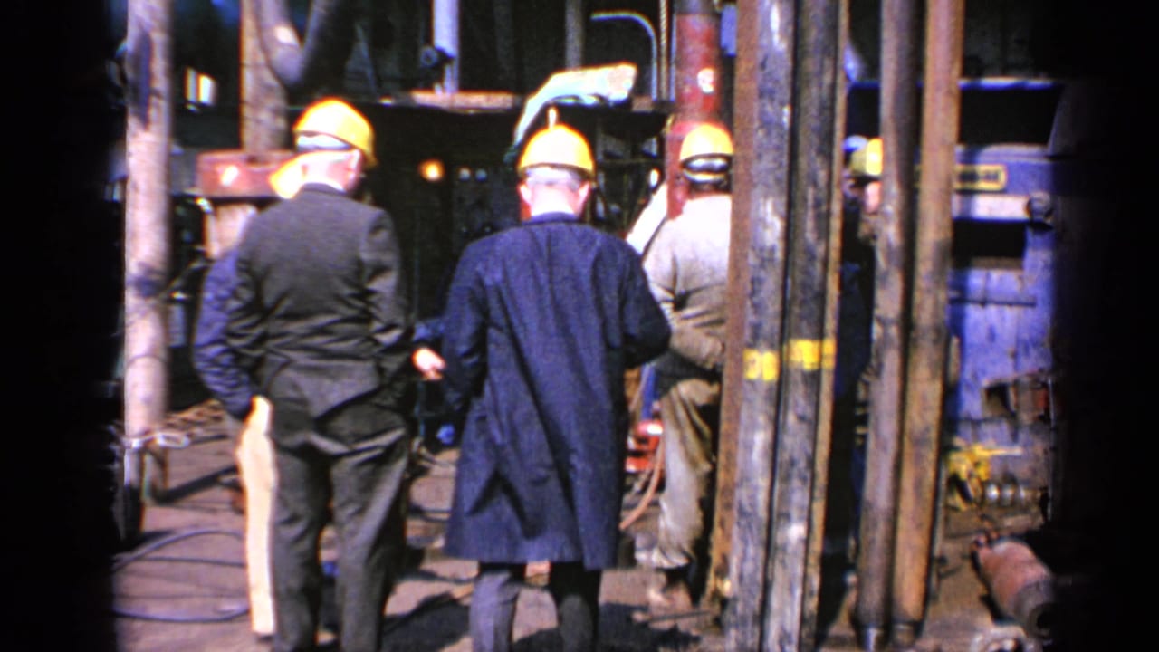 1970 United Kingdom 8mm frame showing workers in yellow hard hats and suited men gathered around heavy industrial equipment.