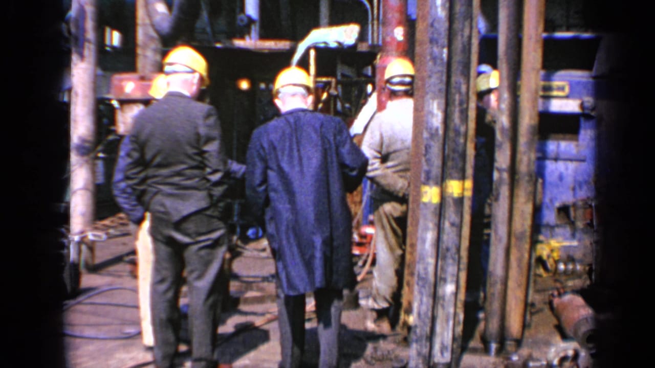 Hard-hatted workers and suited men face machinery among pipes and metal structures.