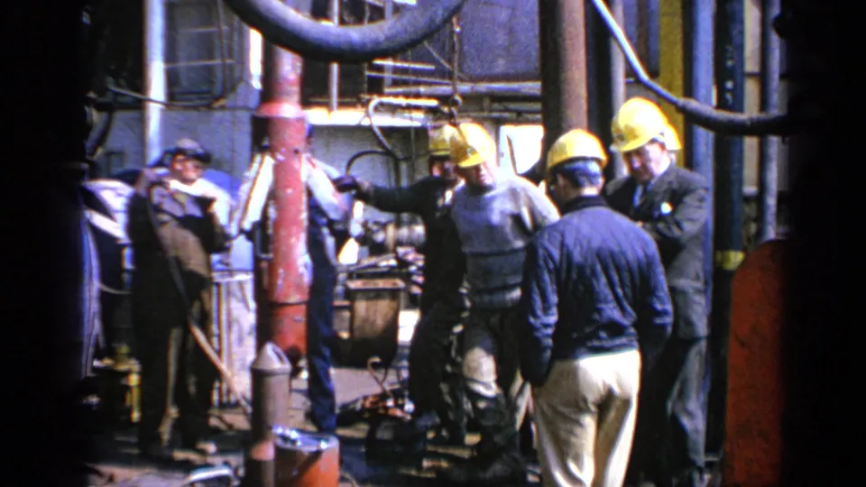 Hard-hatted industrial workers and suited observers gather around machinery on a British factory or plant floor in 1970 footage.