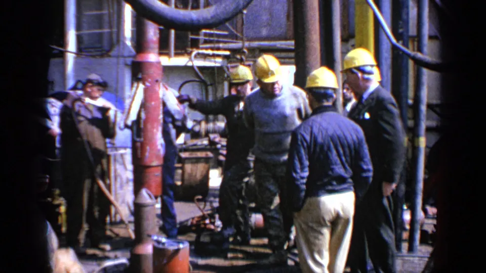 A worker looks toward the camera while others gather near machinery in 1970 United Kingdom industrial home-movie footage.
