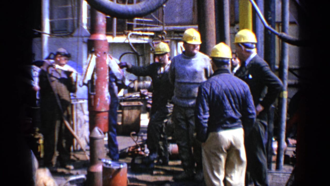 Workers in yellow hard hats stand around industrial machinery and pipes.