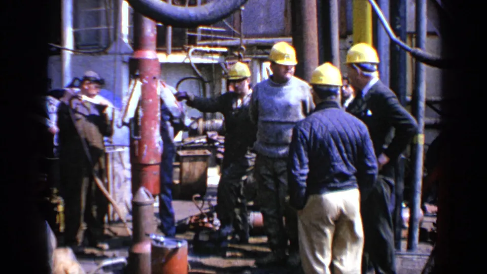 1970 United Kingdom 8mm frame showing workers in hard hats gathered near pipes, valves, and heavy machinery at an industrial site.
