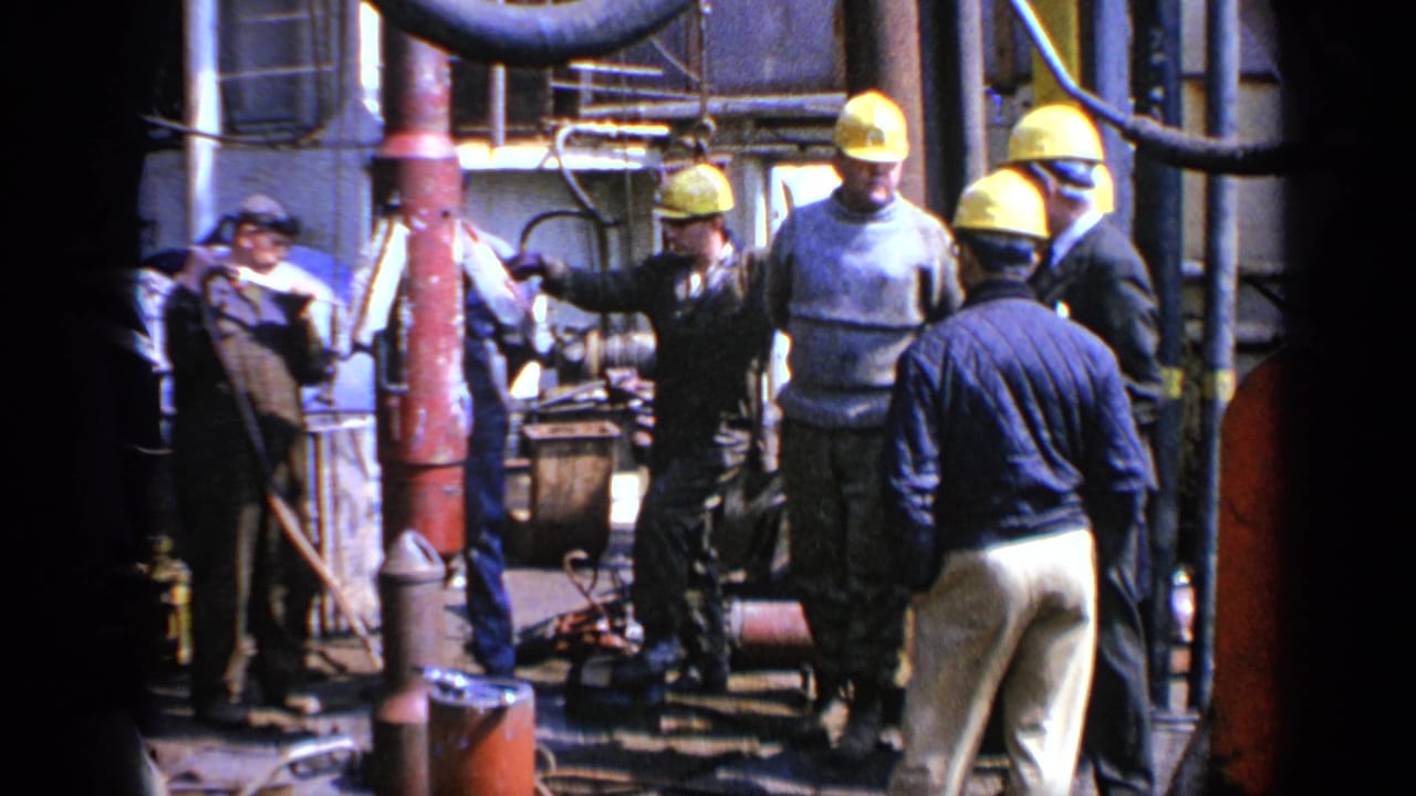 Hard-hatted workers and a suited man stand beside pipes and equipment.