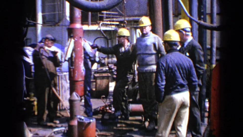 Hard-hatted workers at a 1970 United Kingdom industrial construction setting, seen among red pipework, hoses, and equipment.