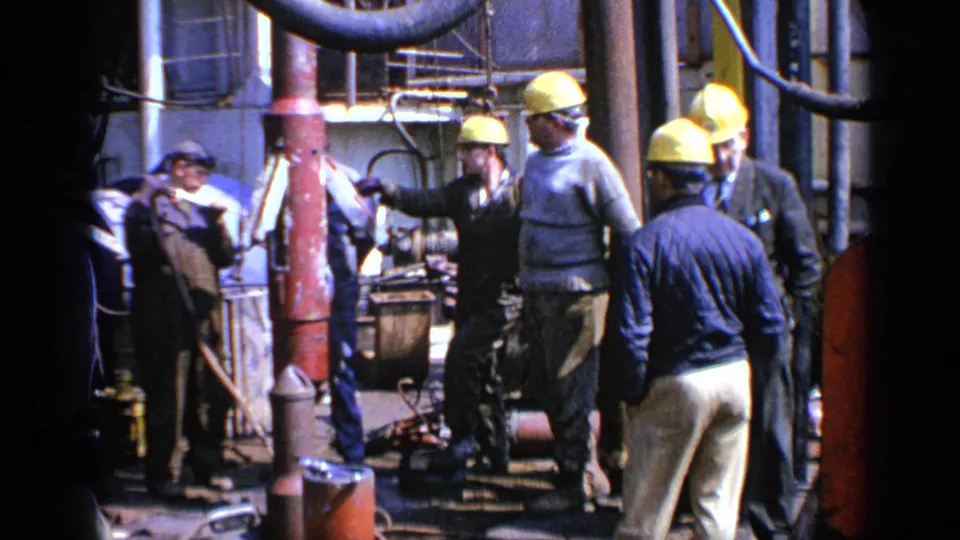 1970 United Kingdom 8mm footage frame of workers beside machinery, hoses, and vertical pipes during an apparent industrial site visit.