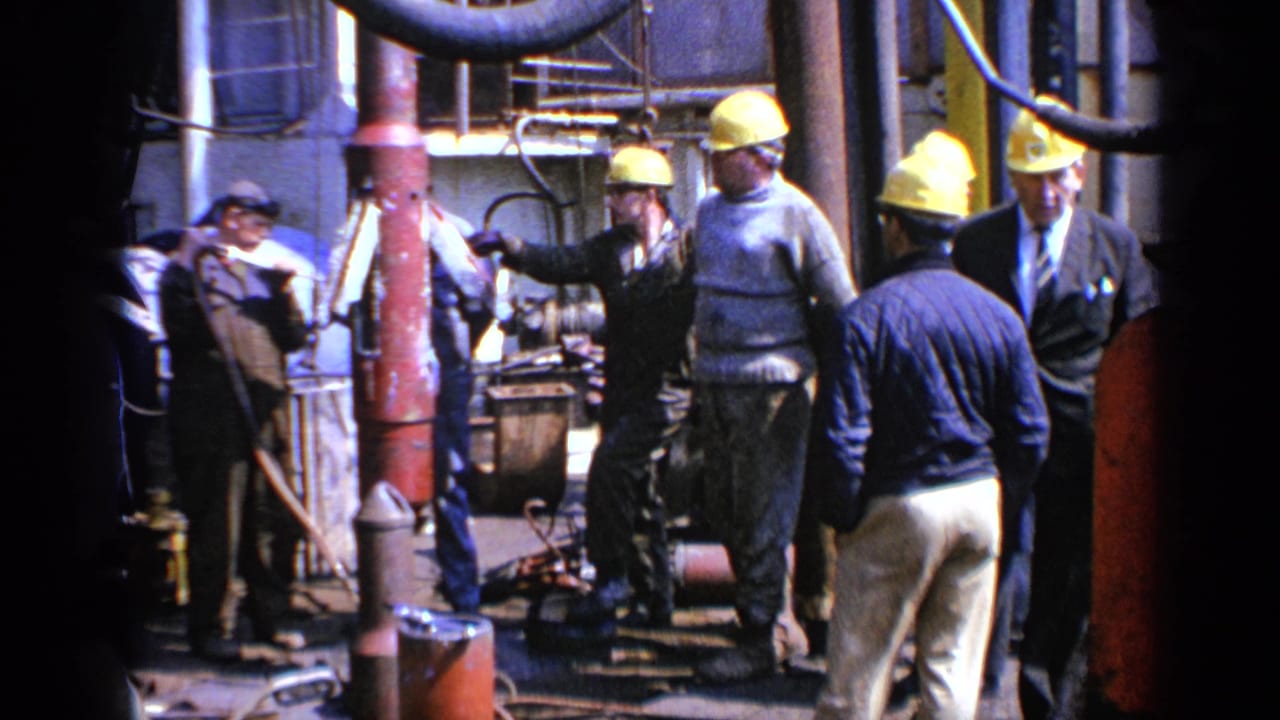 1970 United Kingdom 8mm frame showing men in yellow hard hats standing around heavy machinery and pipes in an industrial setting.