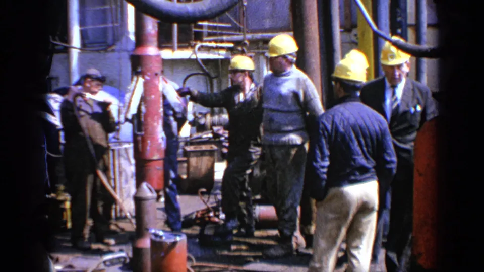 1970 United Kingdom 8mm frame showing men in yellow hard hats standing around heavy machinery and pipes in an industrial setting.