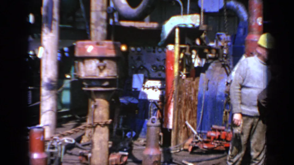 1970 United Kingdom 8mm frame with a worker in a yellow hard hat standing beside pipes, hoses, and heavy workshop equipment.