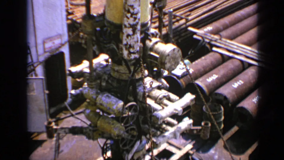 High-angle 1970 United Kingdom frame of pipe bundles, valves, and heavy machinery framed by dark 8mm film borders.