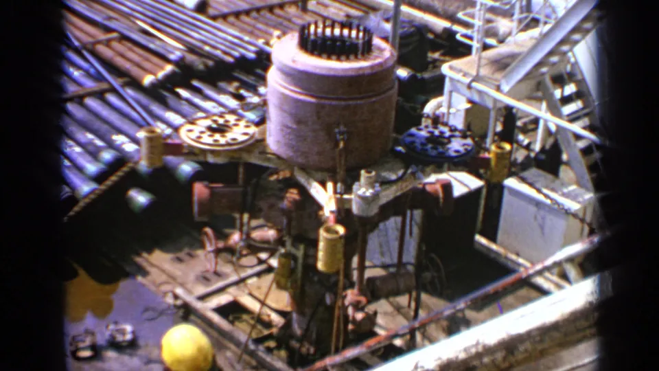 High-angle 1970 United Kingdom frame with cylindrical machinery, pipe stacks, railings, and a yellow hard hat near the lower edge.