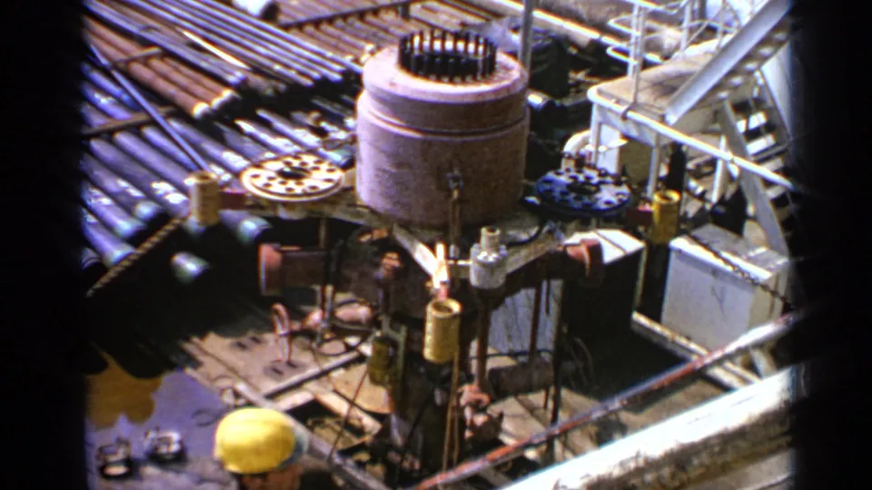 1970 United Kingdom offshore platform view with drilling equipment, bundled pipes, railings, and a hard-hatted worker partly visible below.