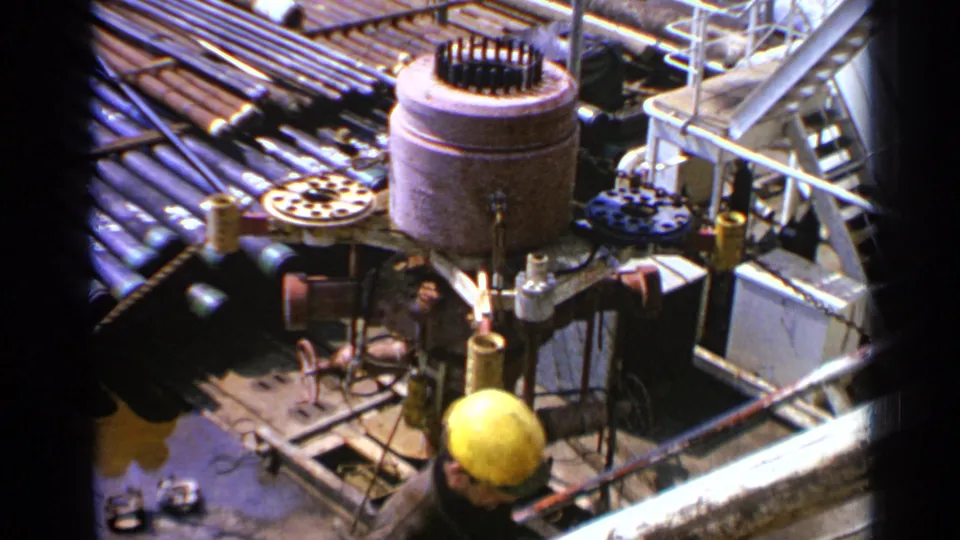 Hard-hatted worker moves beside drilling equipment on a United Kingdom offshore platform, with pipes and metal structures around him.