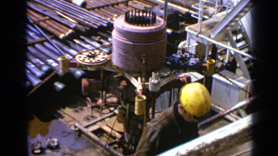 1970 offshore platform frame showing a worker in a yellow hard hat beside drilling equipment, pipes, railings, and deck machinery.