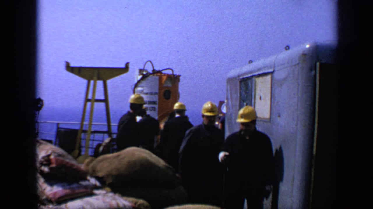 Workers in yellow hard hats move past bags, a trailer, and a white industrial tank near water.