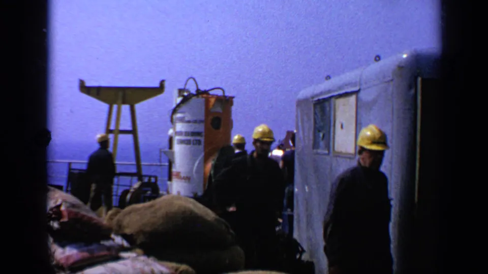 1970 United Kingdom 8mm frame showing industrial workers moving between bags, a trailer, and a white tank beside water.