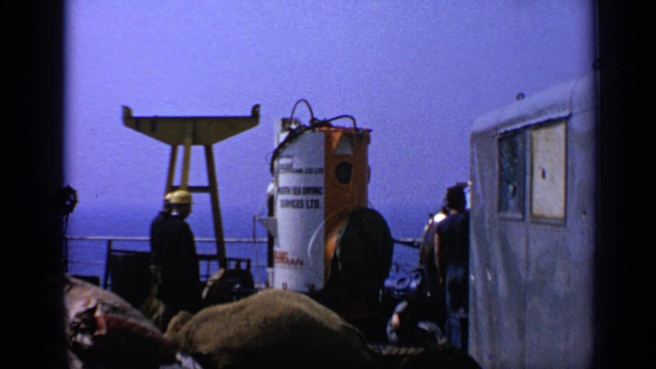 1970 United Kingdom offshore deck scene with crew in hard hats near a labeled diving bell, railings, gear, sea, and blue sky.