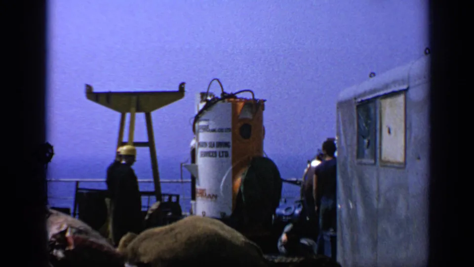 1970 United Kingdom offshore footage shows hard-hatted workers around a white and orange diving bell on a platform deck.
