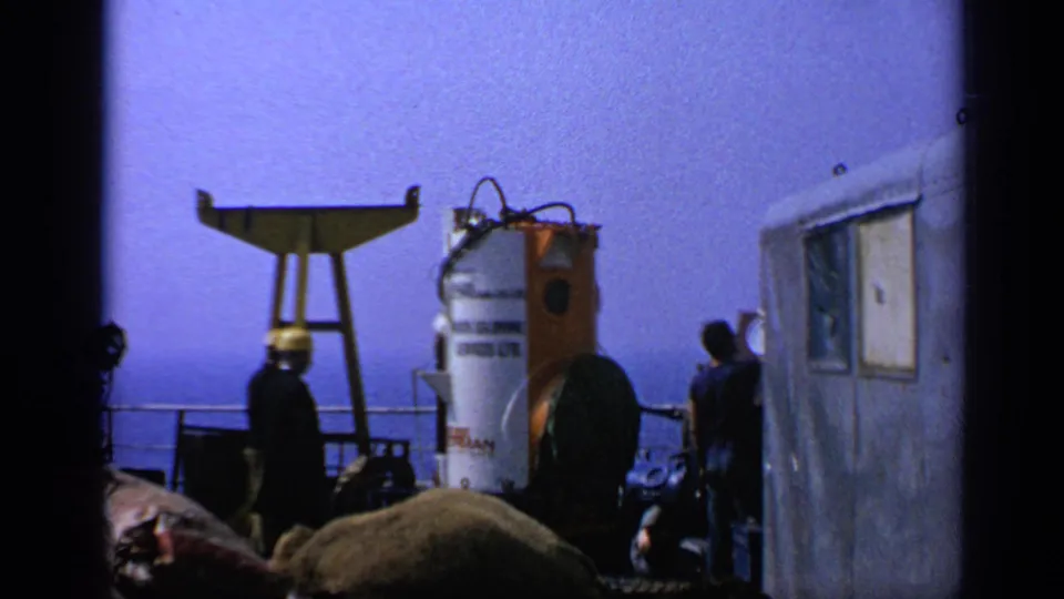 1970 United Kingdom offshore deck view with a silhouetted crew member, diving bell, rig equipment, cabin wall, and sea horizon.