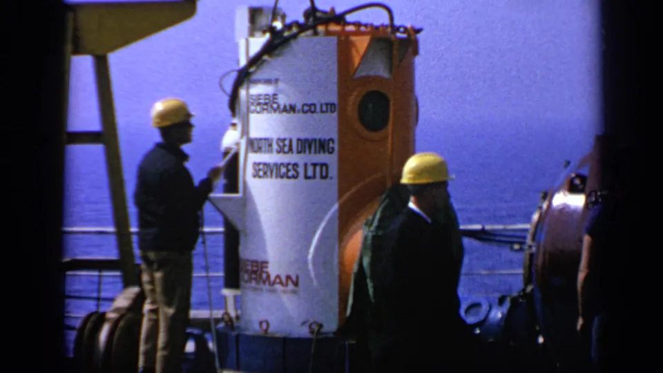 Close 1970 United Kingdom offshore view of workers beside a North Sea Diving Services Ltd. diving bell with sea and railings behind.
