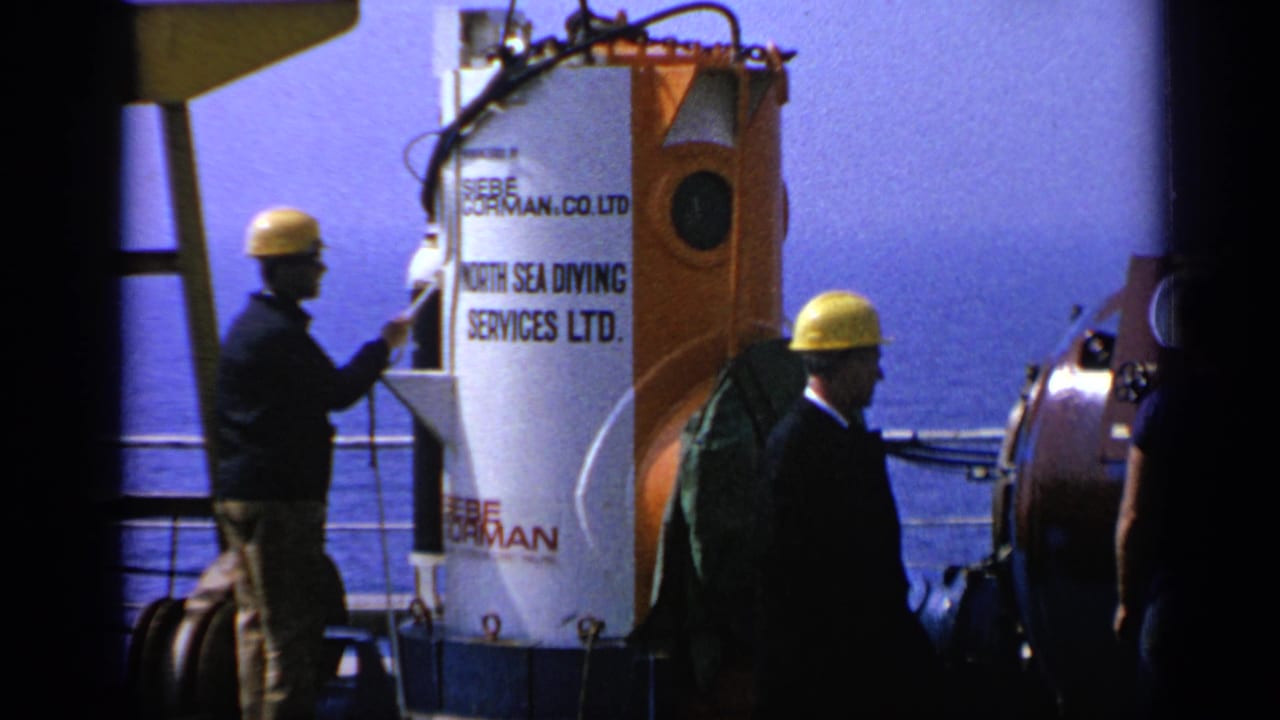 Men in yellow hard hats stand near a white and orange diving chamber on a ship deck.