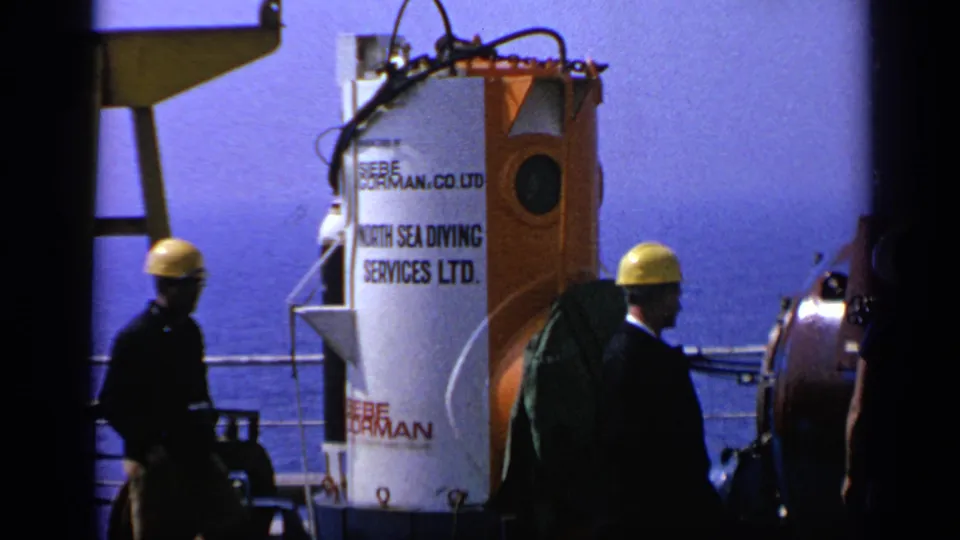 Men in yellow hard hats move past a white and orange diving chamber on a United Kingdom vessel deck in 1970 footage.